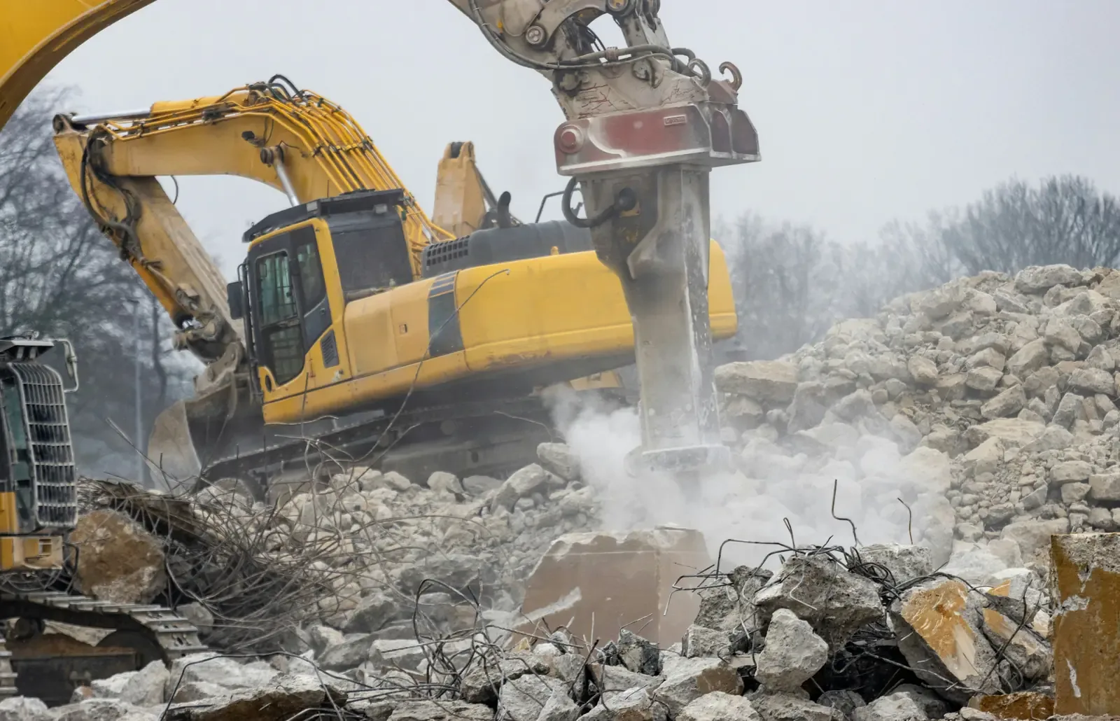 An excavator-mounted hydraulic breaker working a bed of broken concrete, dust in the air