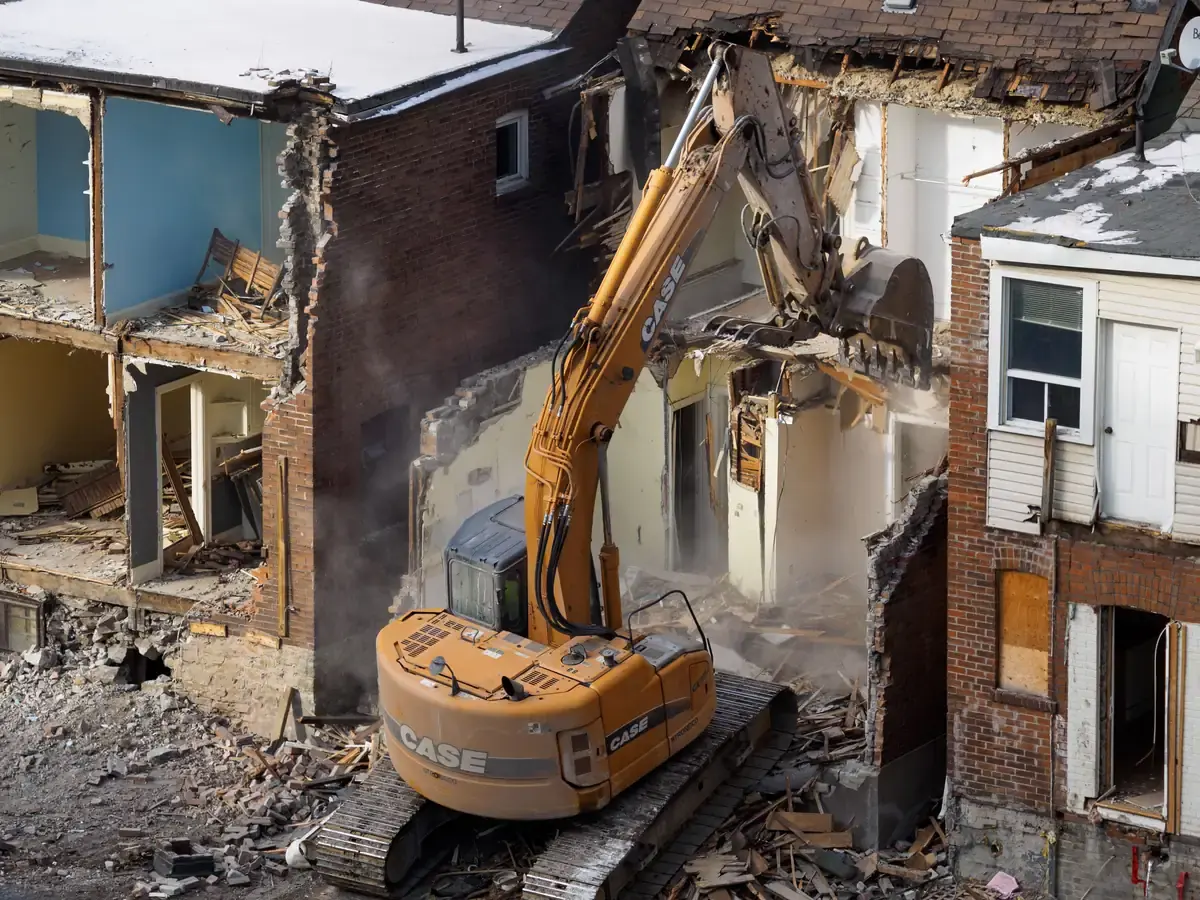 An excavator taking down a brick house, the painted interior rooms open to the air